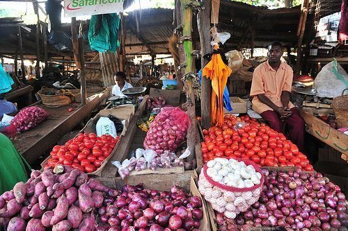 Market stall with piles of red onions, tomatoes, and white bulbs; vendors sit behind the produce.