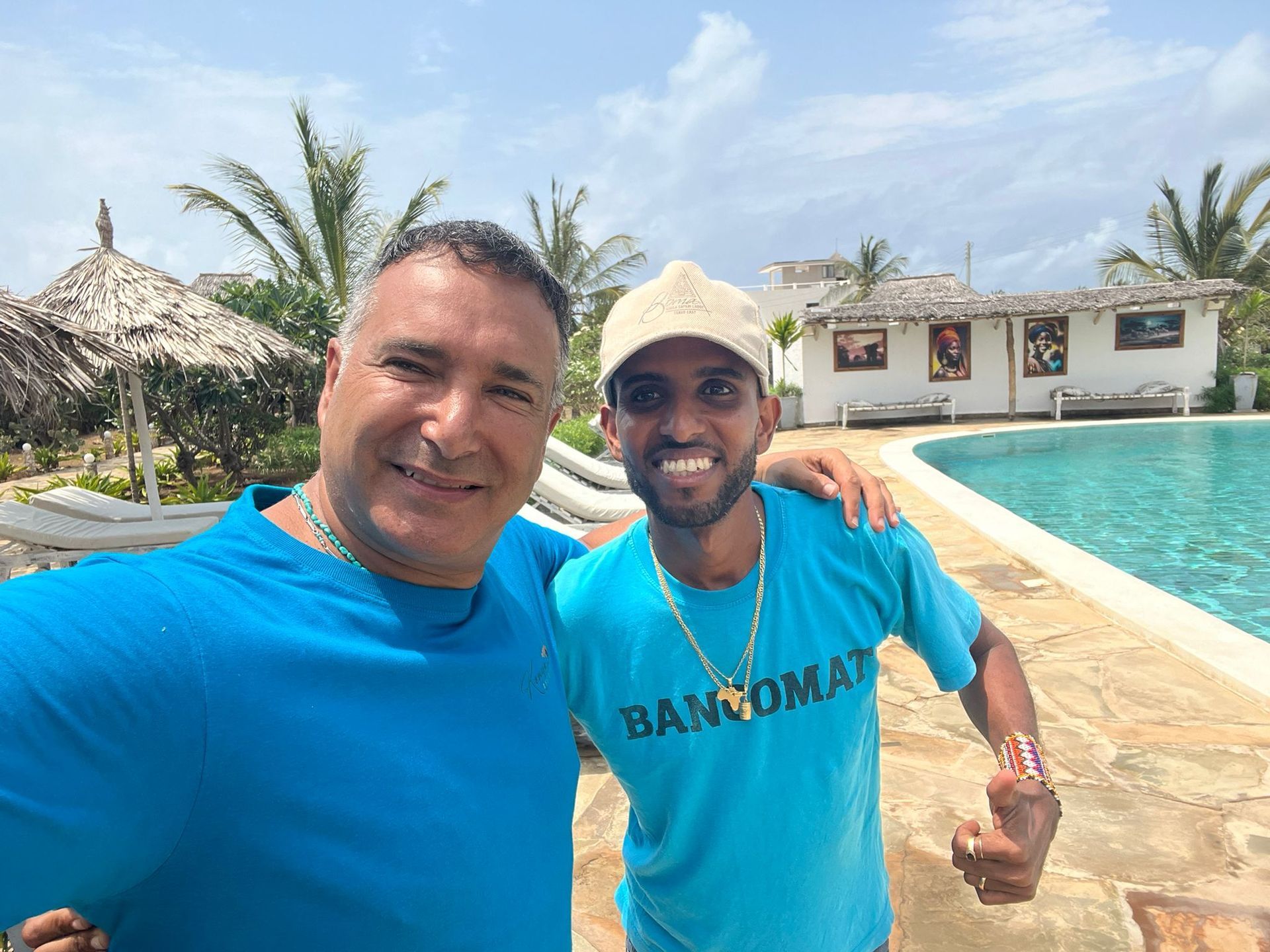 Two men smiling by a pool at a sunny resort, one in a blue shirt and one in a turquoise shirt and cap.