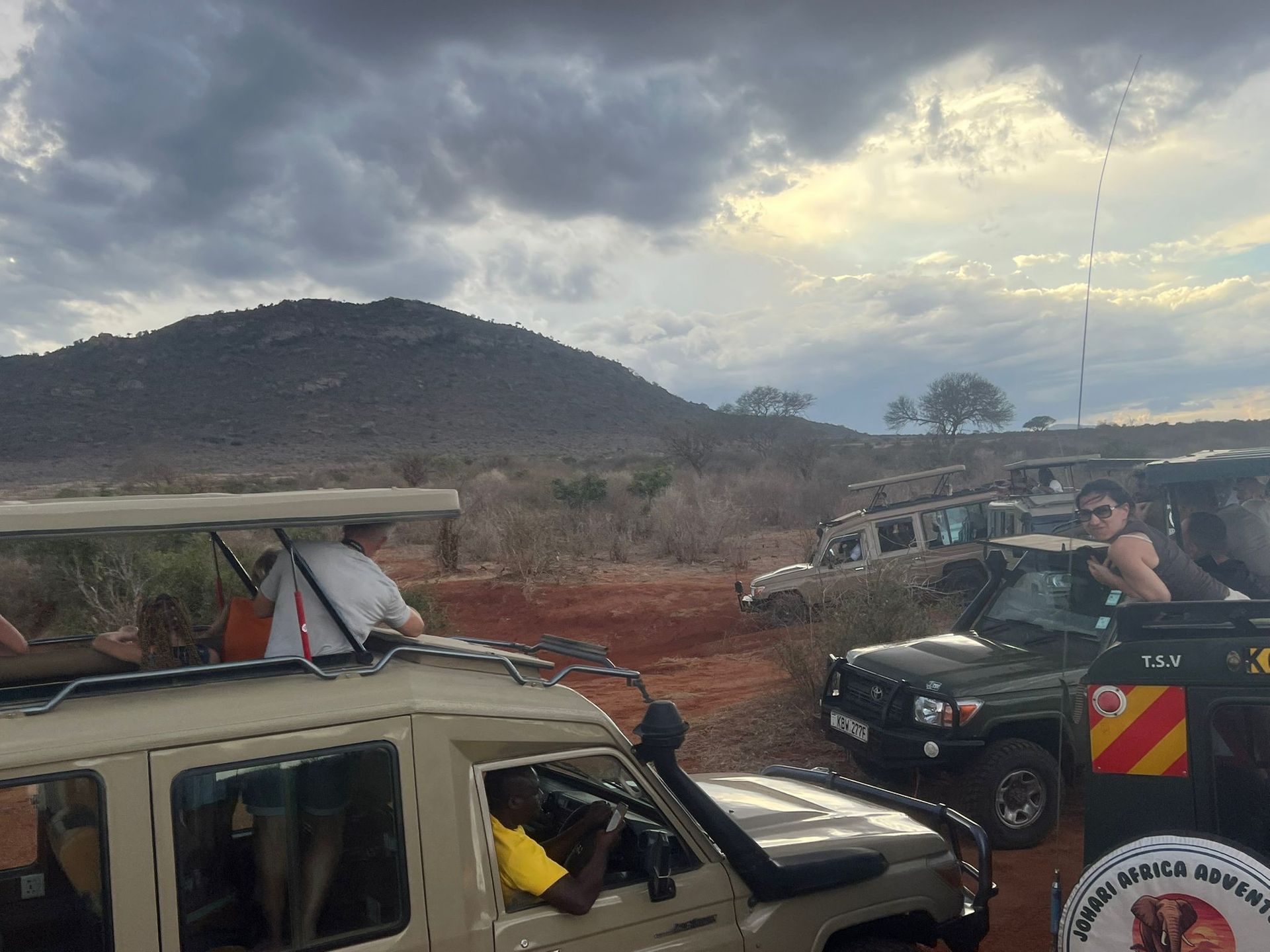 Off-road vehicles on a dirt road under cloudy skies, with people riding in the back near a hillside