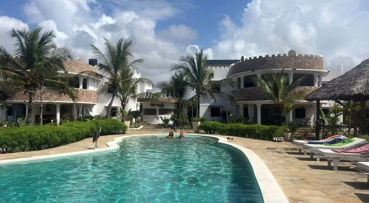 Tropical resort pool with turquoise water, lounge chairs, palm trees, and villa-style buildings under a partly cloudy sky