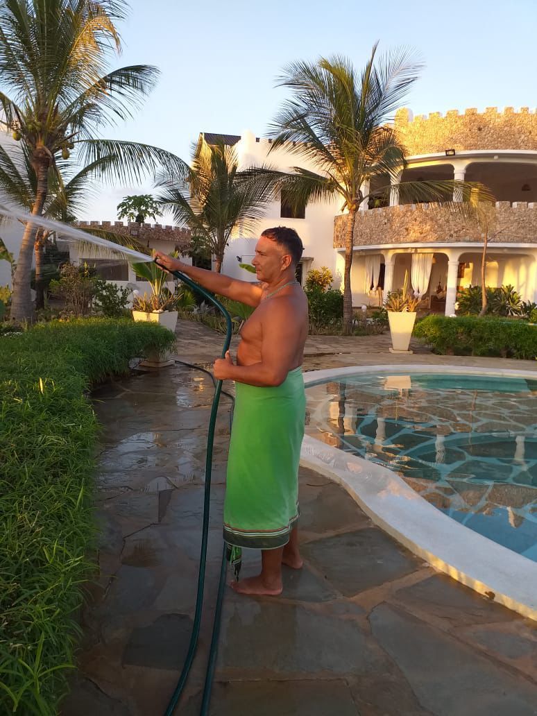 Person in a green wrap stands by a pool, holding a hose in a tropical resort courtyard.