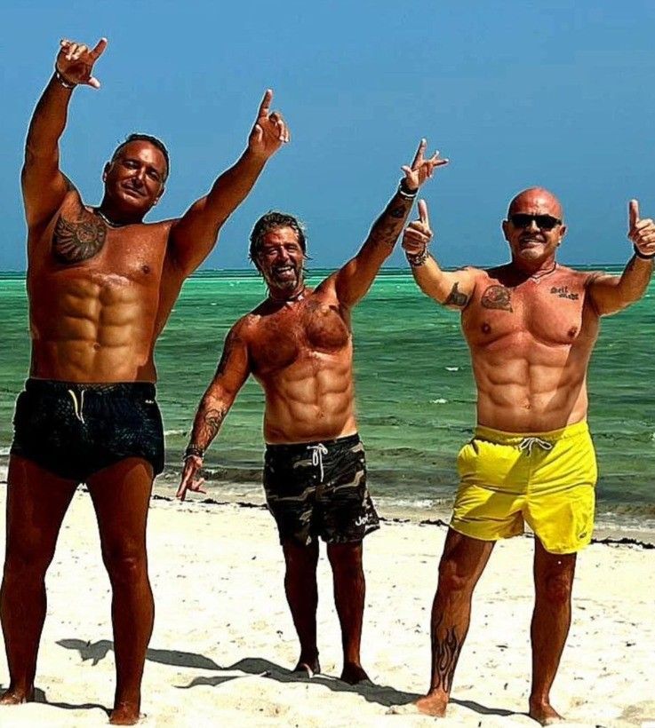 Three shirtless men posing on a sunny beach with arms raised, ocean and blue sky behind them