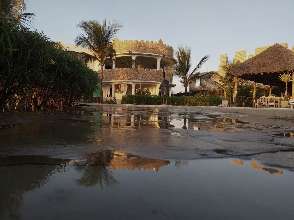 Thatched huts beside a reflective pond at sunset, with palm trees and a tropical lodge in the background