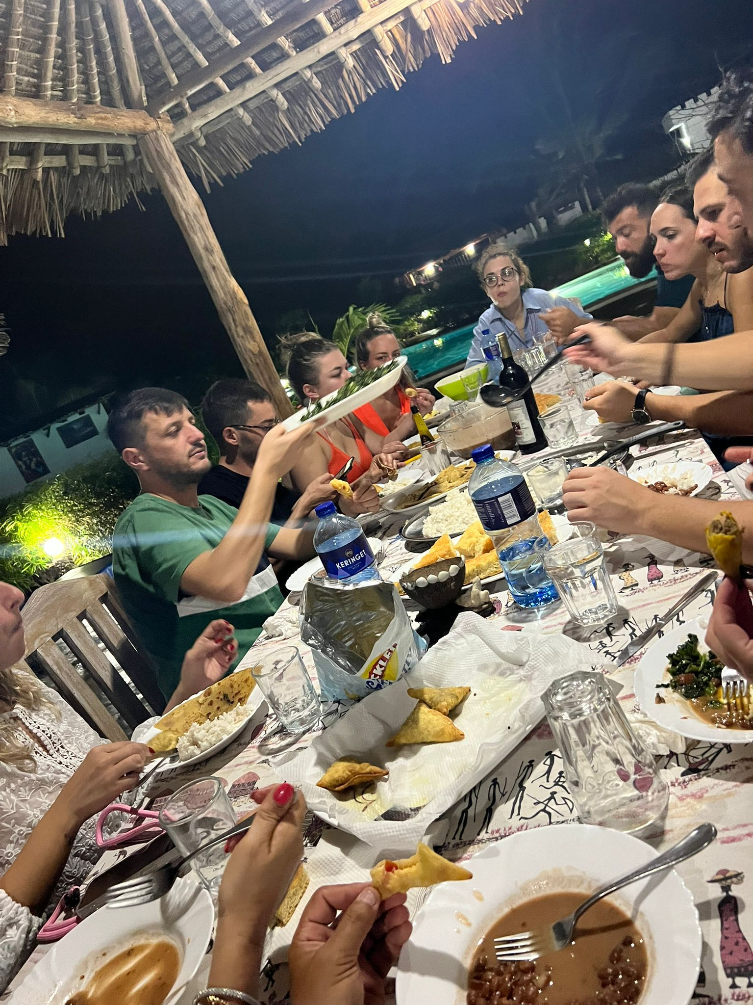 People dining at a long outdoor table under a thatched roof at night, with food, drinks, and lit candles.