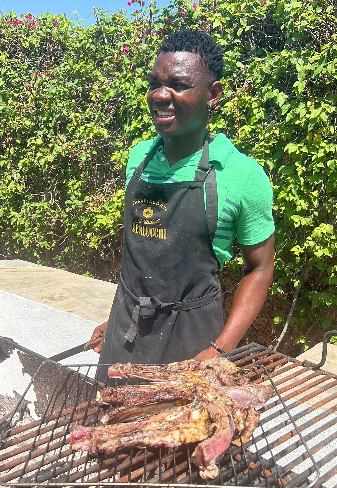 Man grilling meat outdoors in a black apron, smiling beside a barbecue with greenery behind him