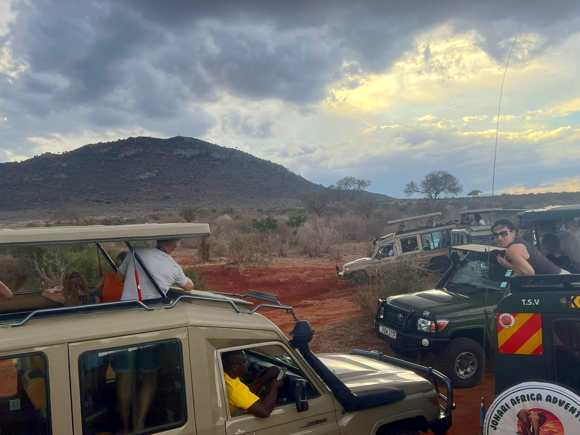 Off-road vehicles on a dirt road near hills under cloudy sky, with people inside and on top of one vehicle