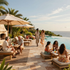 Resort poolside terrace with umbrellas, lounge chairs, and guests dining beside the ocean at sunset