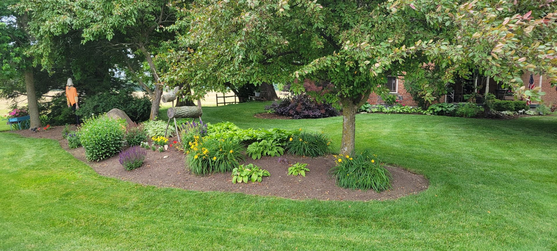 A lush green lawn surrounds a landscaped flower bed with trees and various plants.
