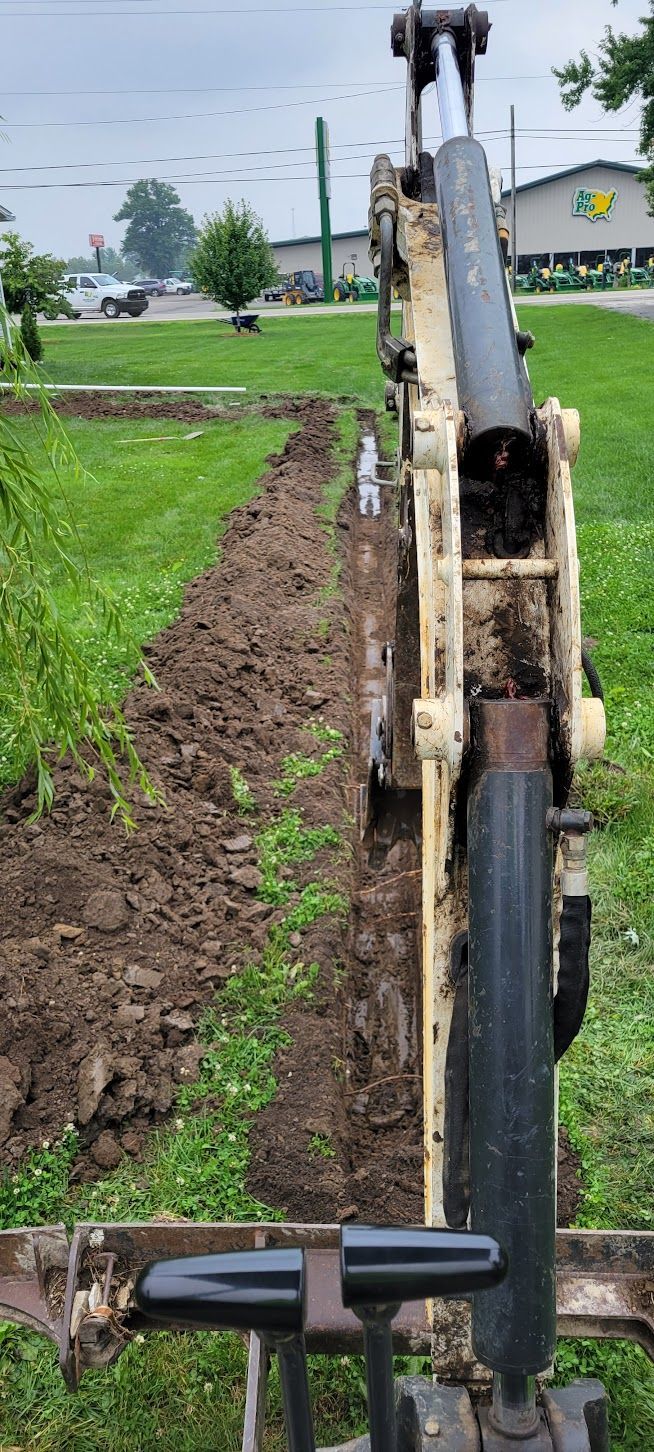 An excavator digging a trench in a grassy area near a road with a building.