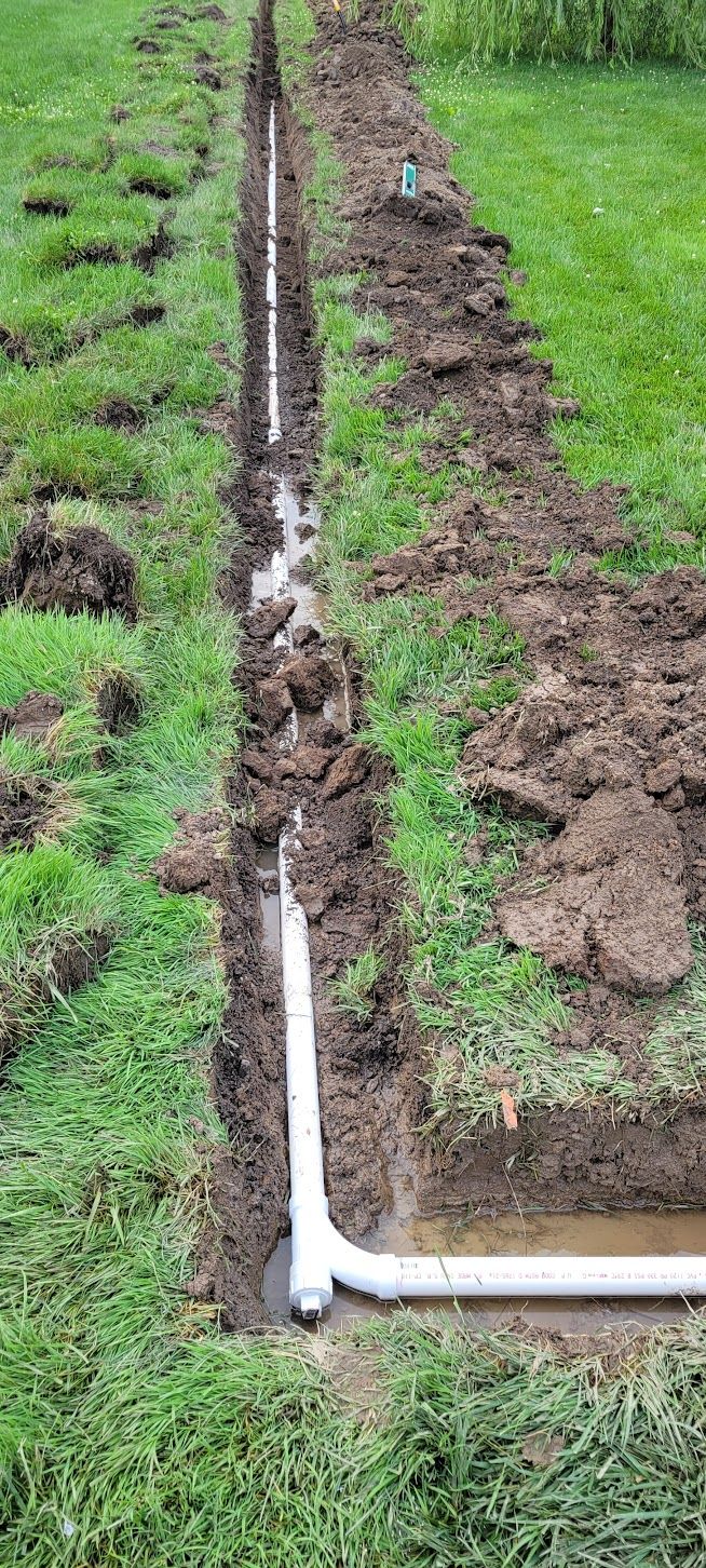 A trench with white PVC pipes laid inside. Dirt is piled up beside the trench in a grassy area.