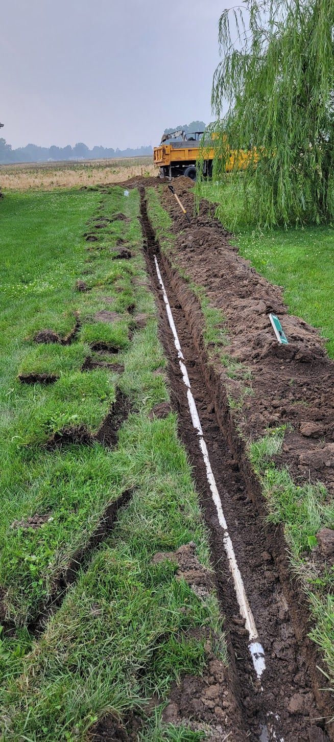 A trench is dug in a grassy area, a white pipe laid in it. A yellow vehicle is in the distance.