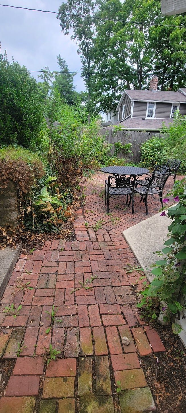 Brick pathway leading to a patio with black metal furniture, surrounded by greenery and a house.