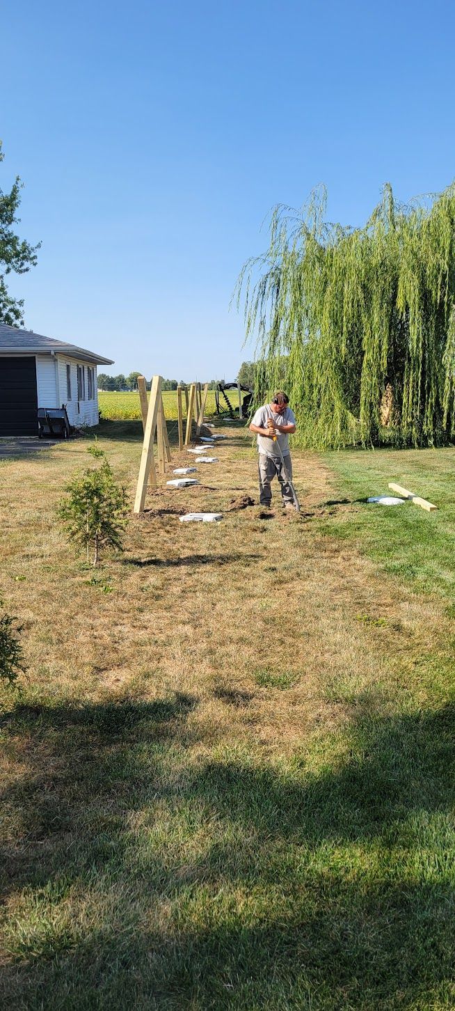 Person working on a fence in a yard with wooden posts and a house in the background on a sunny day.