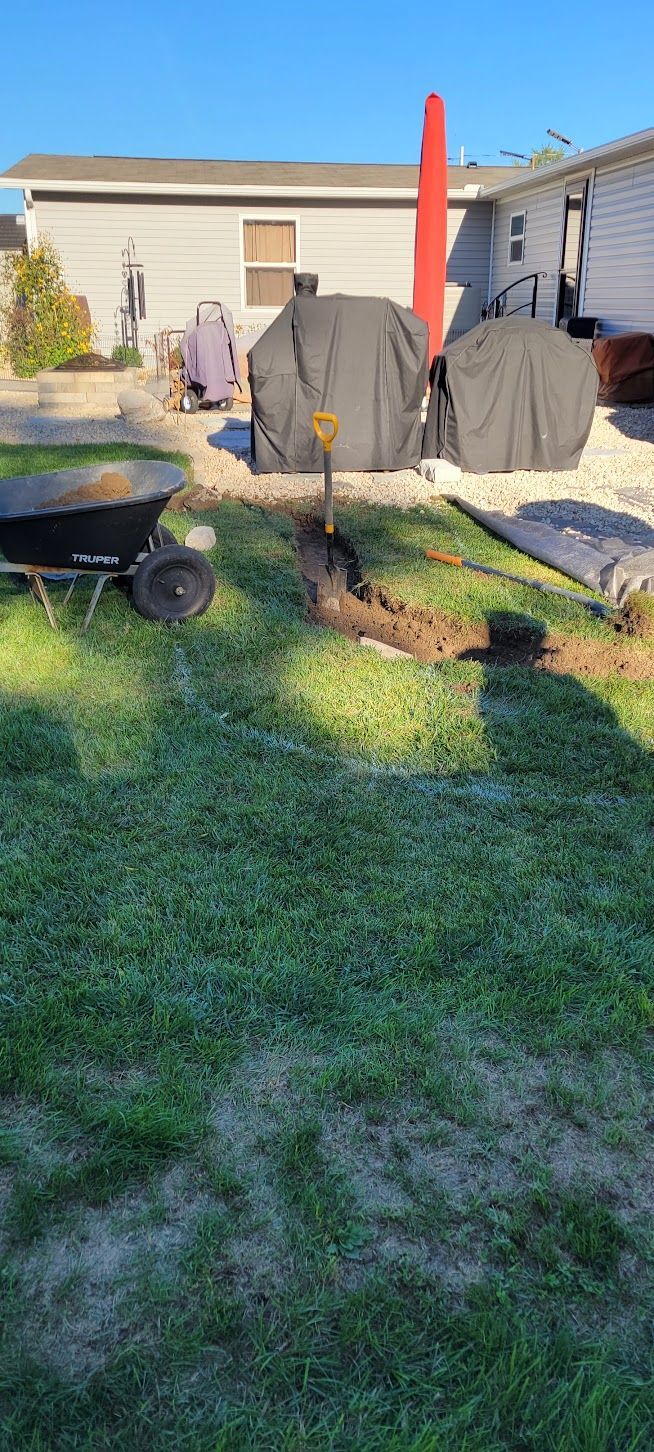 A backyard with a trench dug in the grass, wheelbarrow, covered grills, and a house in the background.