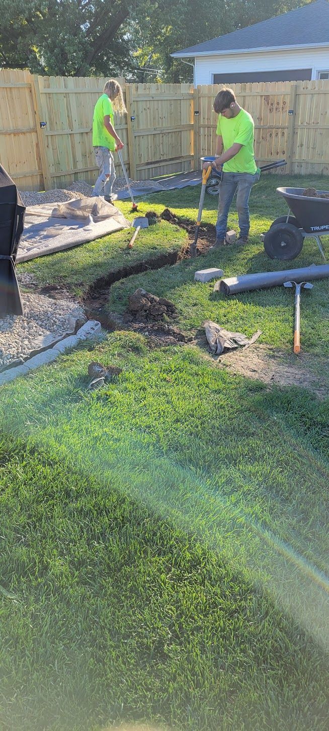 Two workers digging trench in yard, next to a fence. One using a shovel, the other with a pickaxe.