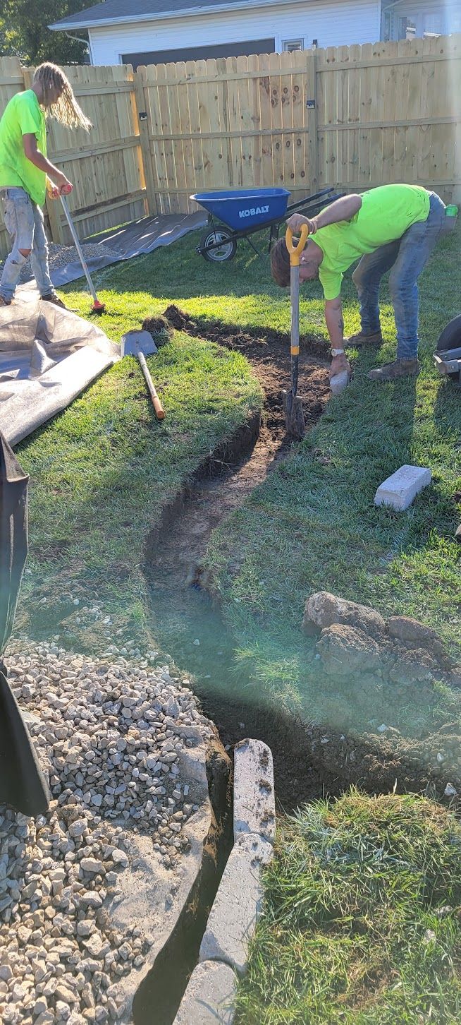 Two workers digging a trench in a grassy yard, near a stone border and a fence.