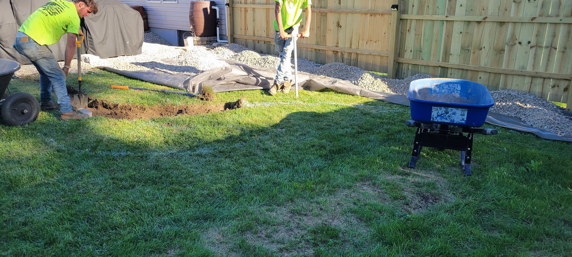 Man digging in a yard near a rock bed and a blue fertilizer spreader.