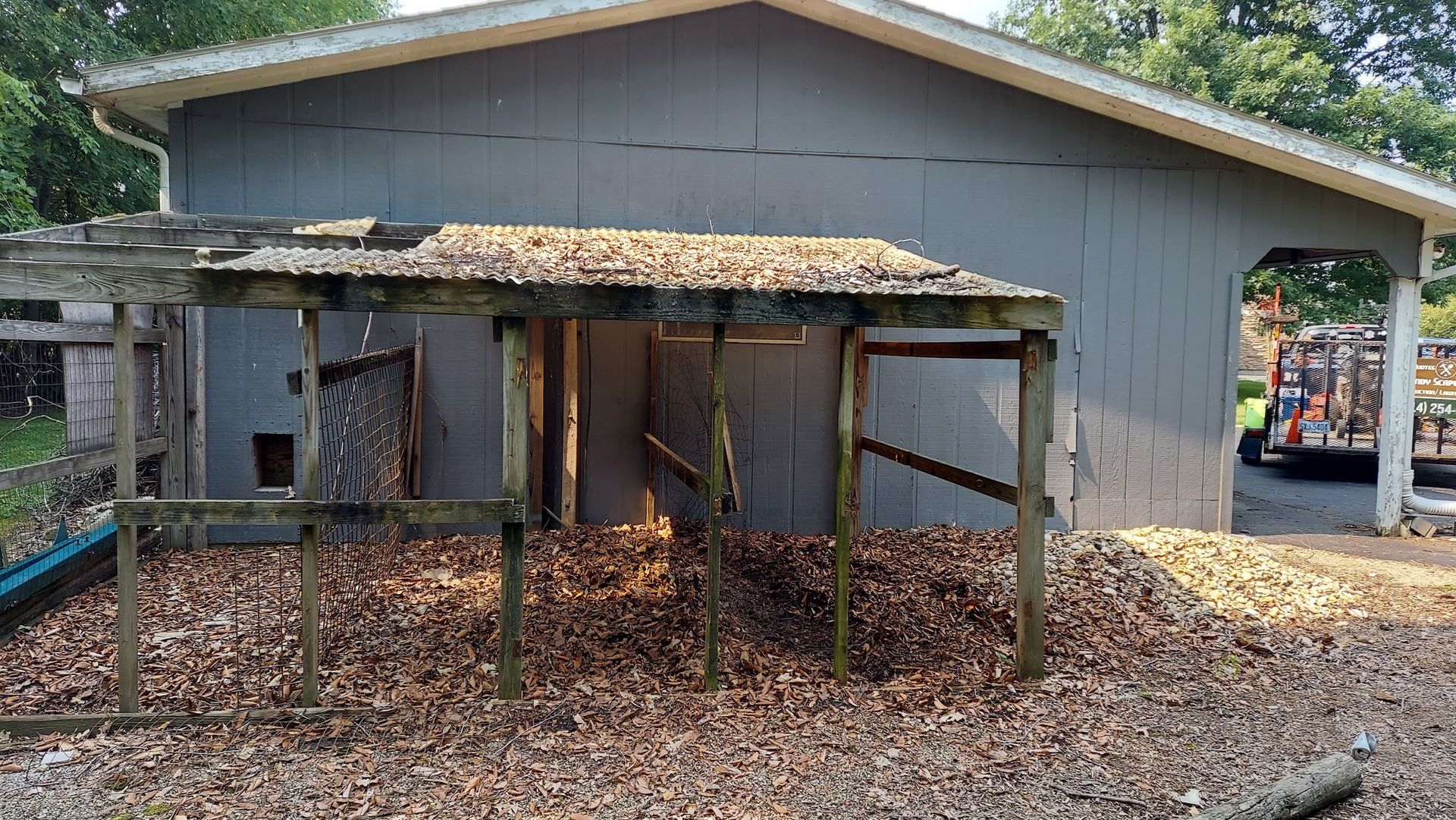 Dilapidated wooden structure with debris-covered roof and ground, set against a gray building with a carport.