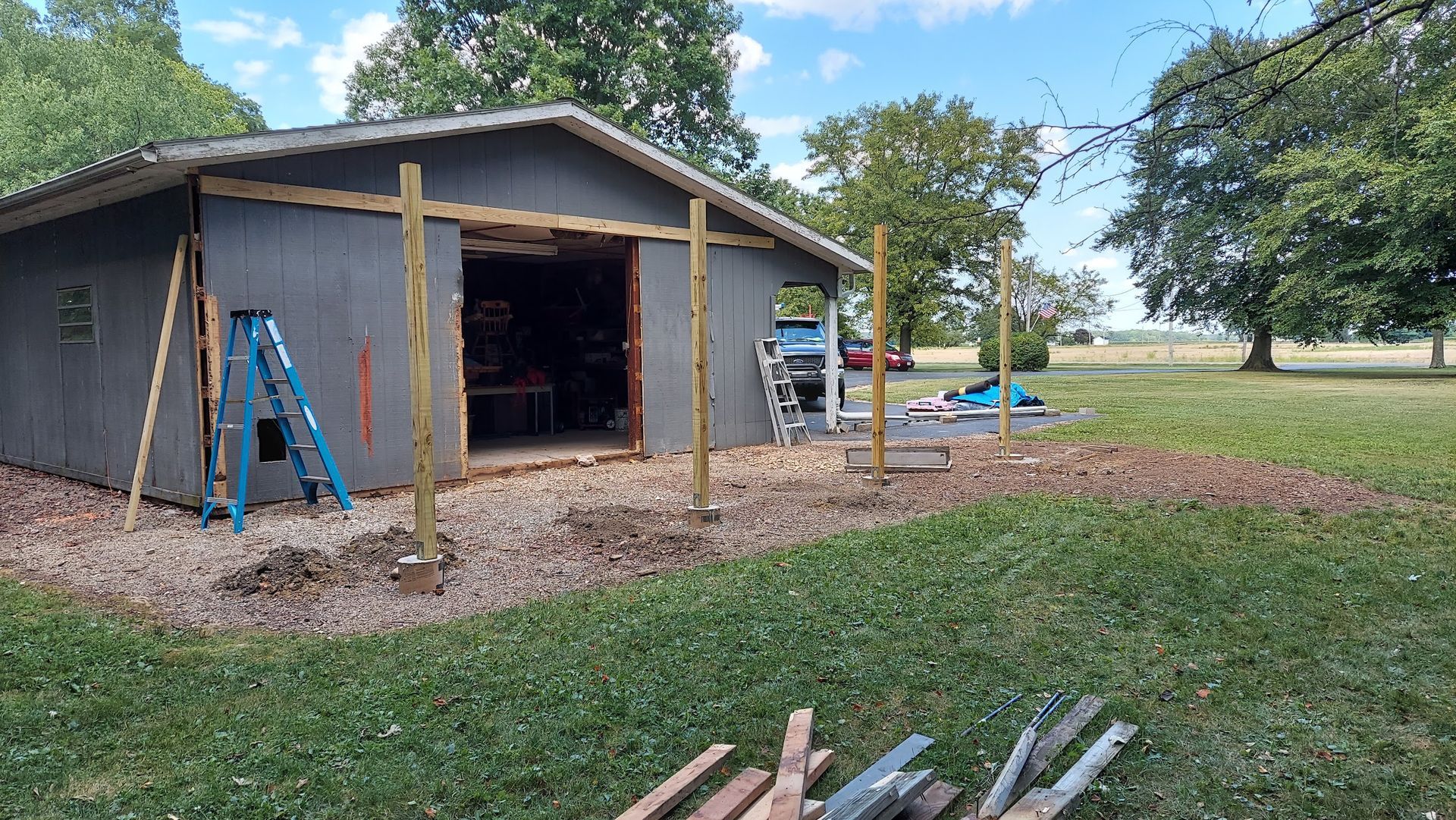 A shed with an open doorway, being expanded with wooden posts; blue ladder and construction materials in a grassy yard.