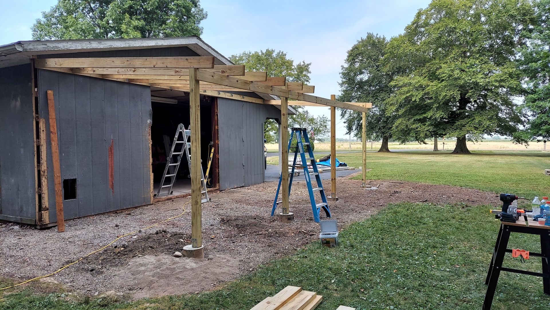 A partially constructed wooden pergola attached to a gray shed in a grassy yard.
