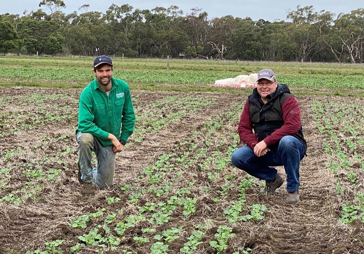 Two people kneel in a muddy field with young crops, one in a green jacket and one in a black vest.