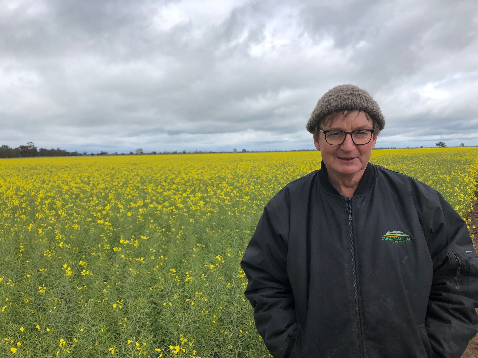 Person in a black jacket and knit hat standing in a yellow flower field under cloudy skies.