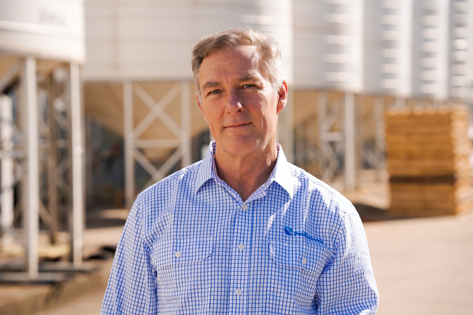 Man standing outdoors in a blue checkered shirt at an industrial site with storage tanks in the background