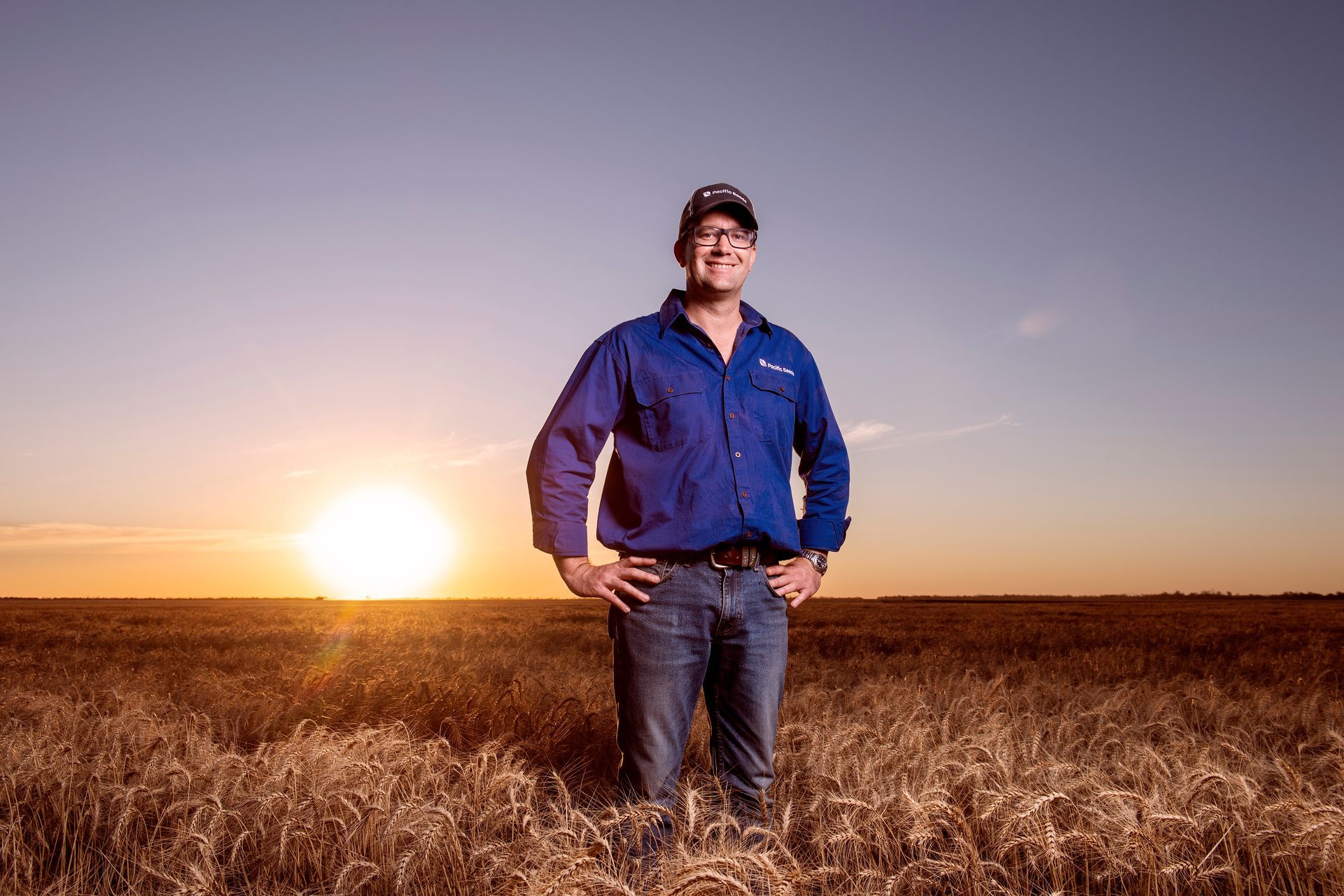 Man standing in a field at sunset, wearing a blue shirt and jeans, with golden sky behind him