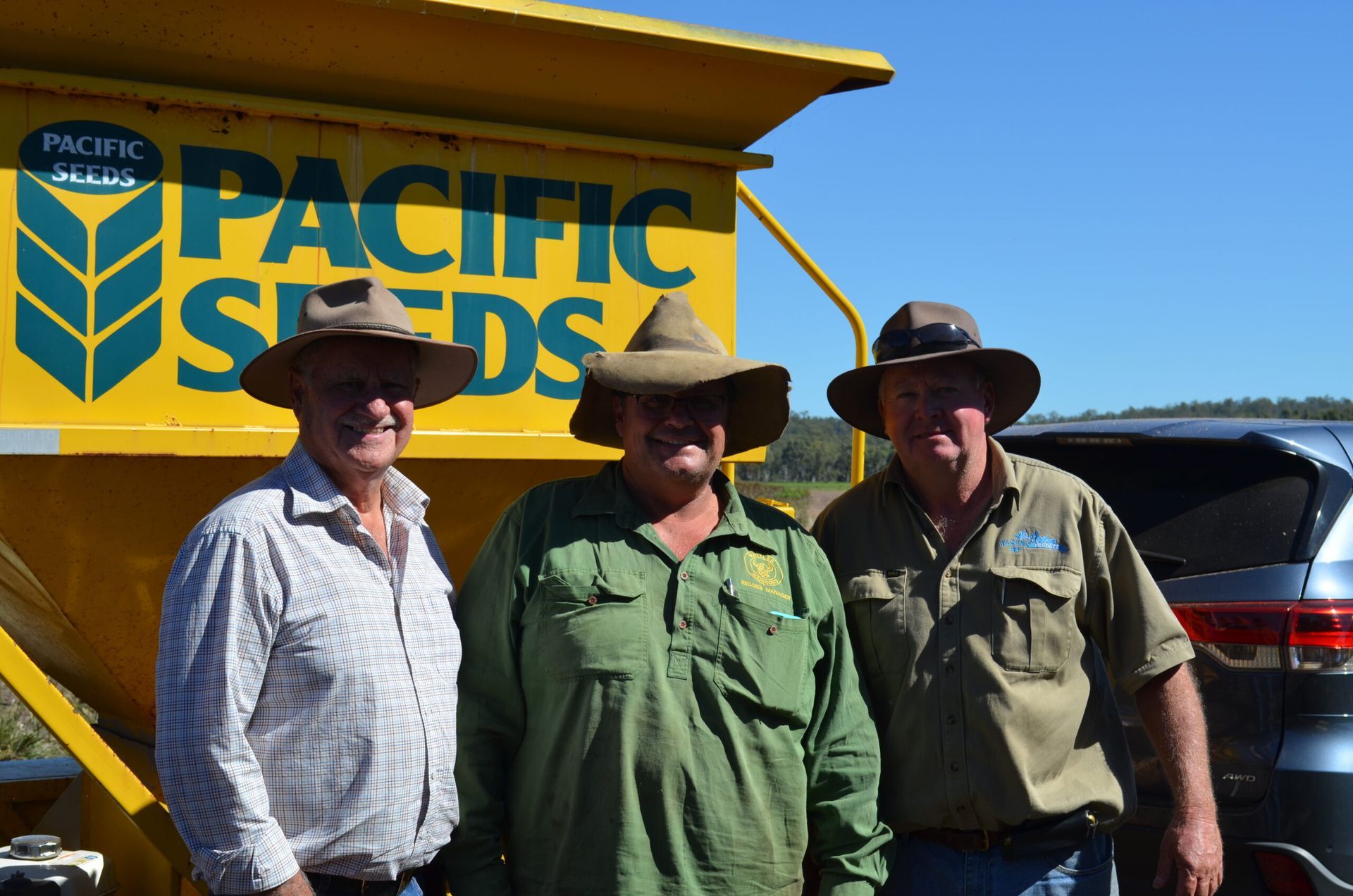 Three people in cowboy hats standing beside a yellow Pacific Seeds truck under a clear blue sky