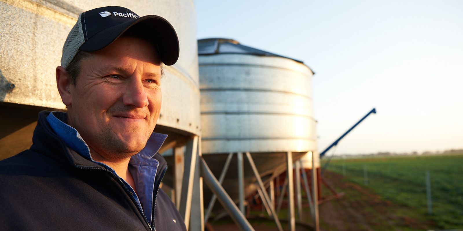 Man in a cap standing beside a grain silo in a field at sunset