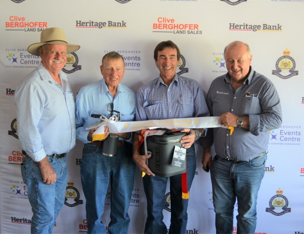 Four men hold a long fish at a fishing event with sponsor banners in the background.