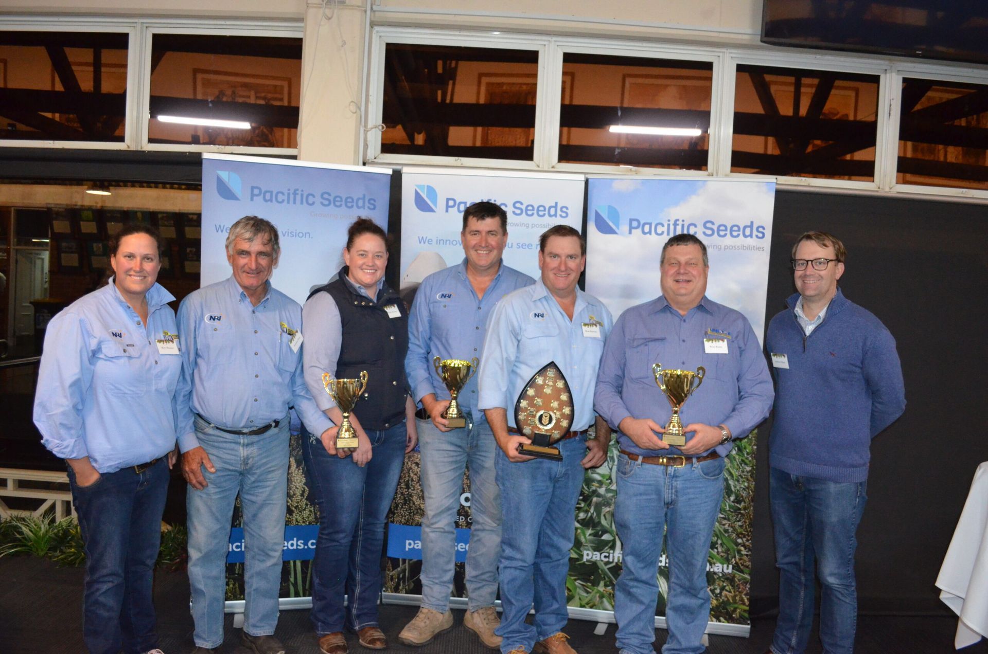Group of seven people in blue shirts holding trophies and awards at a podium backdrop