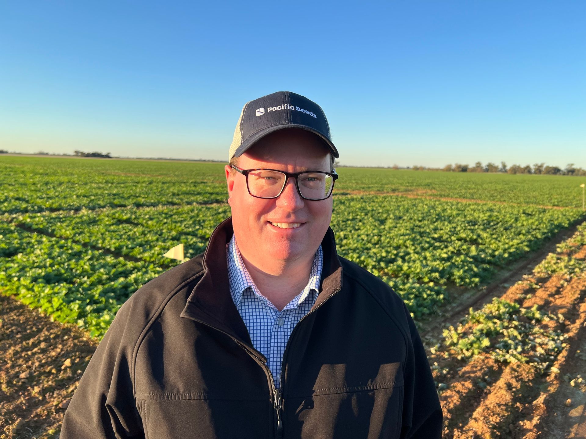 Person in a black jacket and cap smiling in a green field under a clear blue sky.