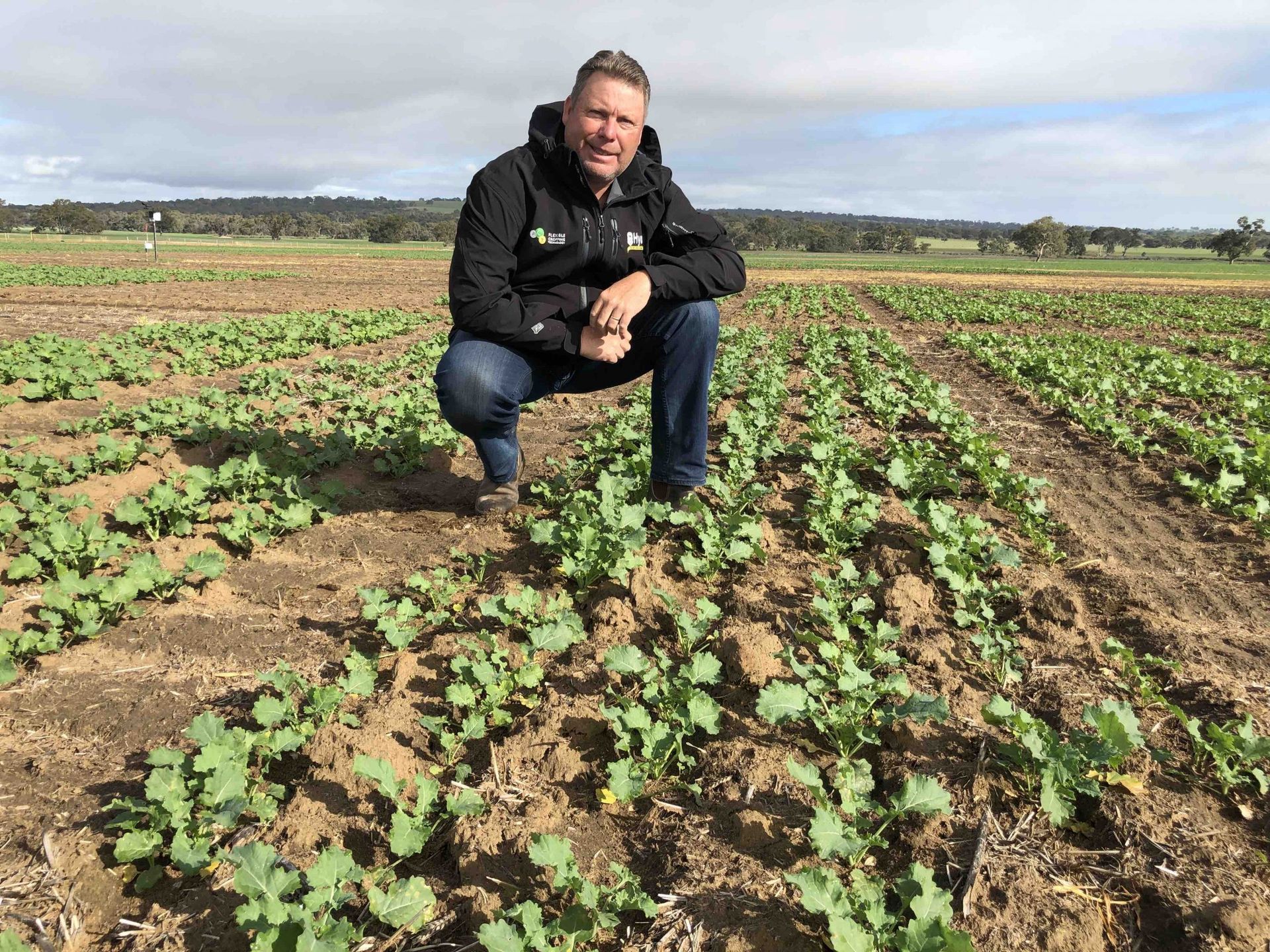 Person crouching in a field of young green crops, with rows of soil stretching into the distance.