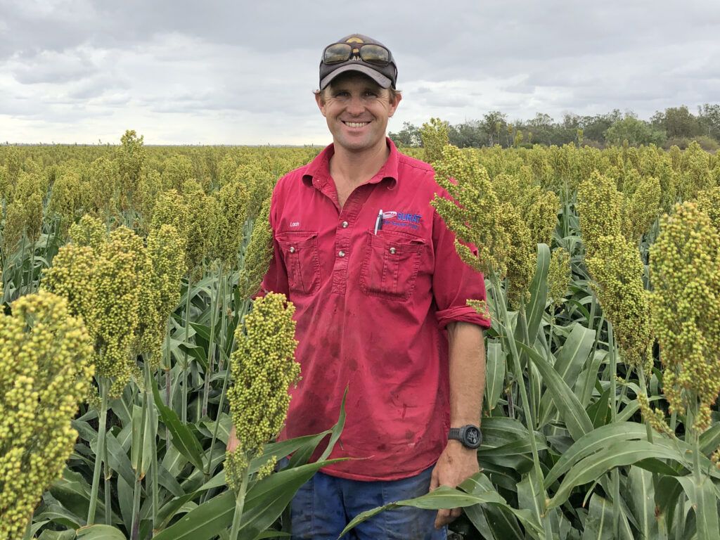 Smiling person in red shirt standing in a crop field with tall green plants under cloudy sky