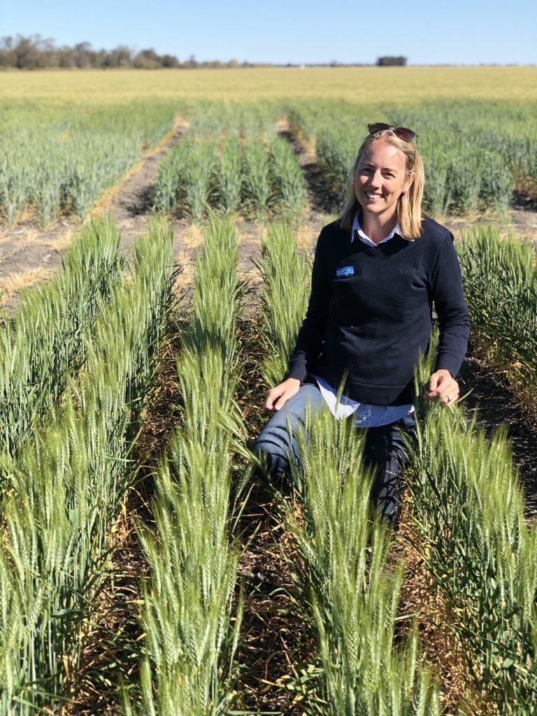 Woman kneeling in green crop rows in a sunny field, smiling at the camera.