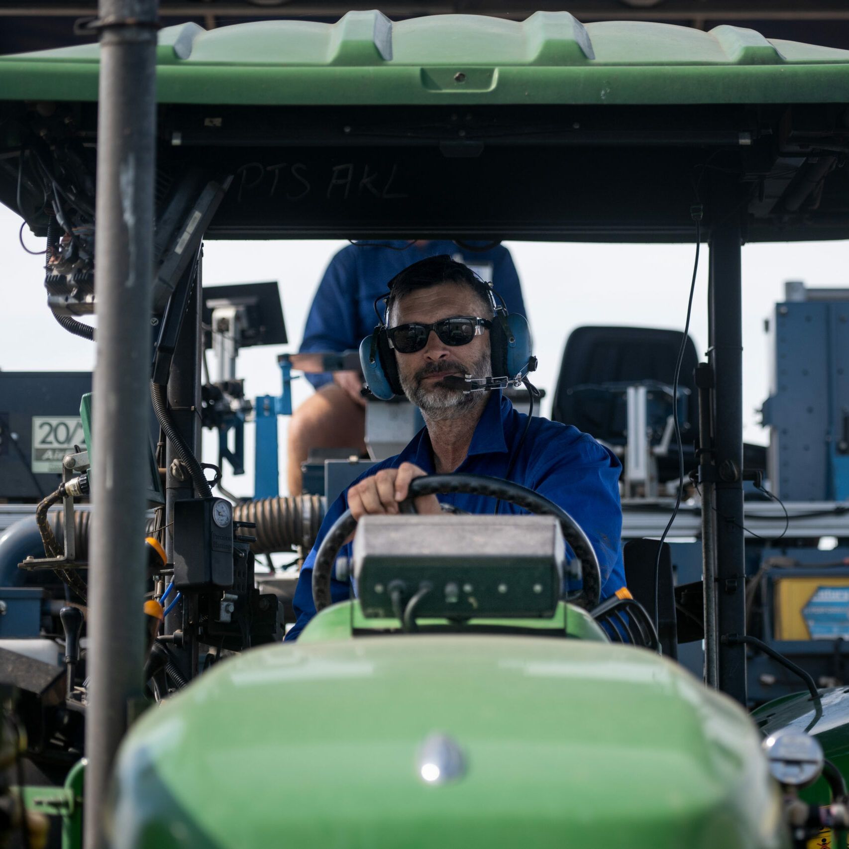 Man in blue shirt and headset driving a green tractor, viewed from the front cabin.