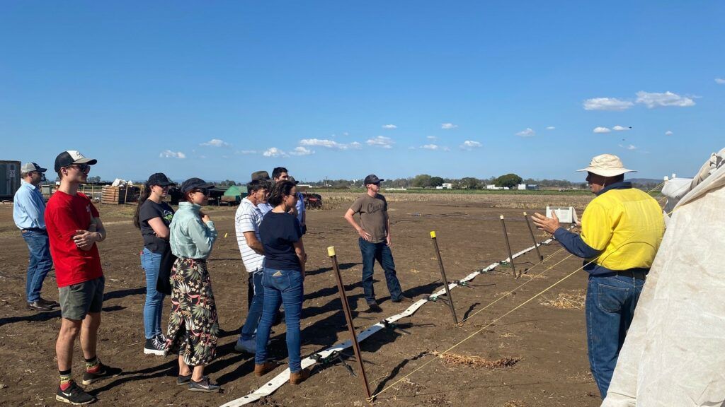 People gathered outdoors near a field, listening to a person in a yellow shirt speaking.