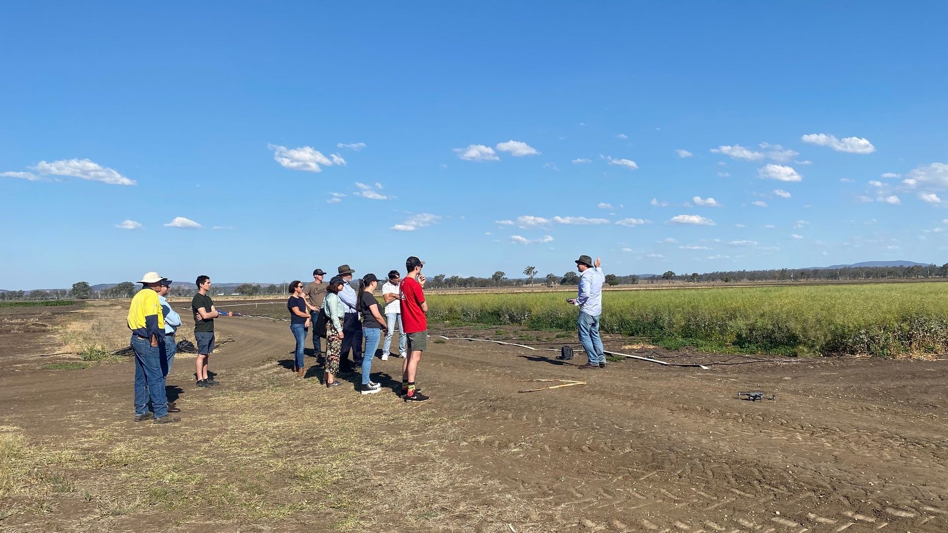 Group of people standing in a field under a bright blue sky, gathered near a dirt road.