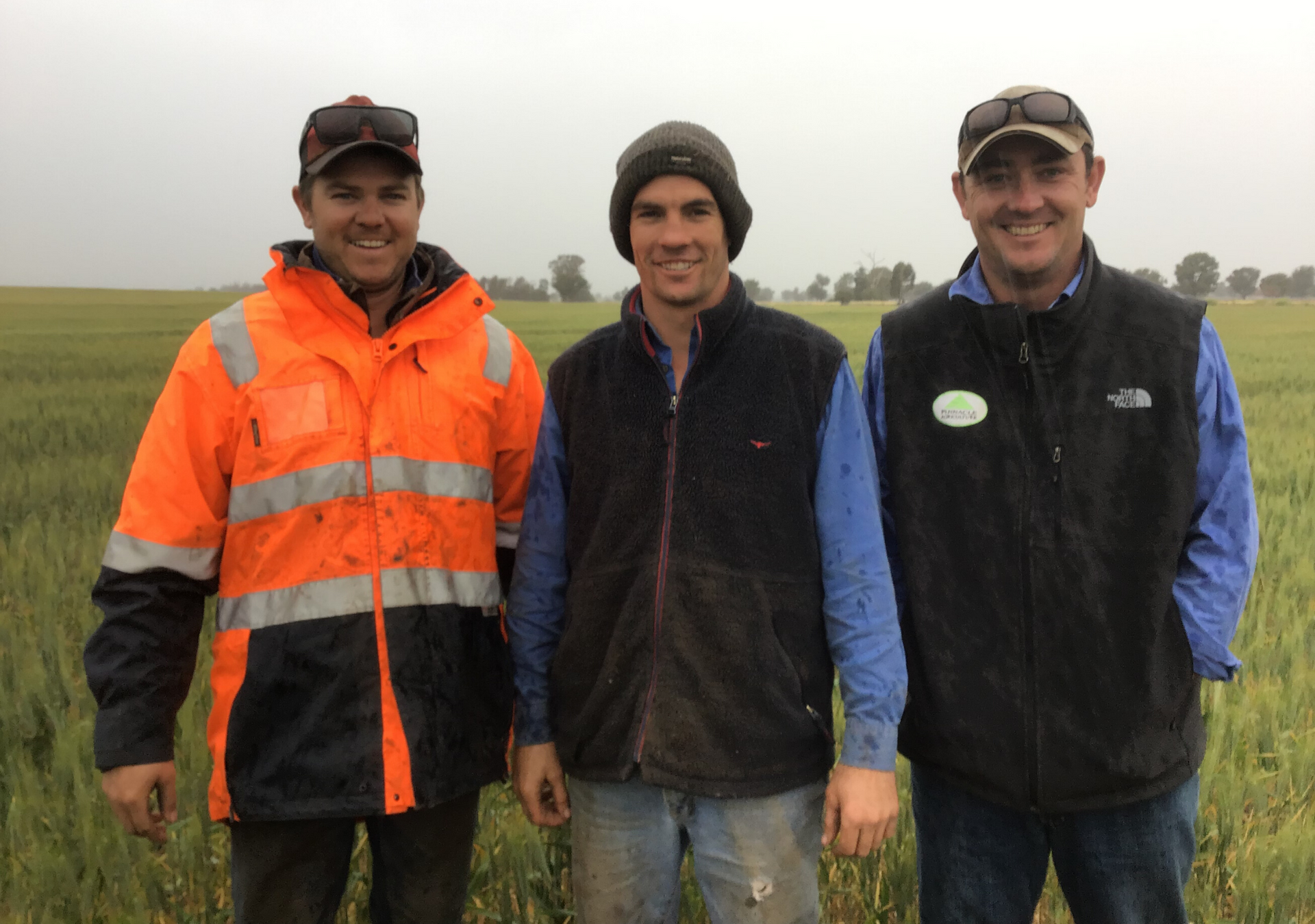 Three people standing in a grassy field, one in a bright orange jacket, under an overcast sky