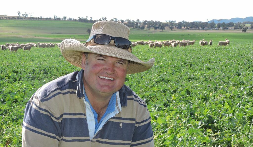 Smiling person in a straw hat standing in a green field with crops and livestock in the background