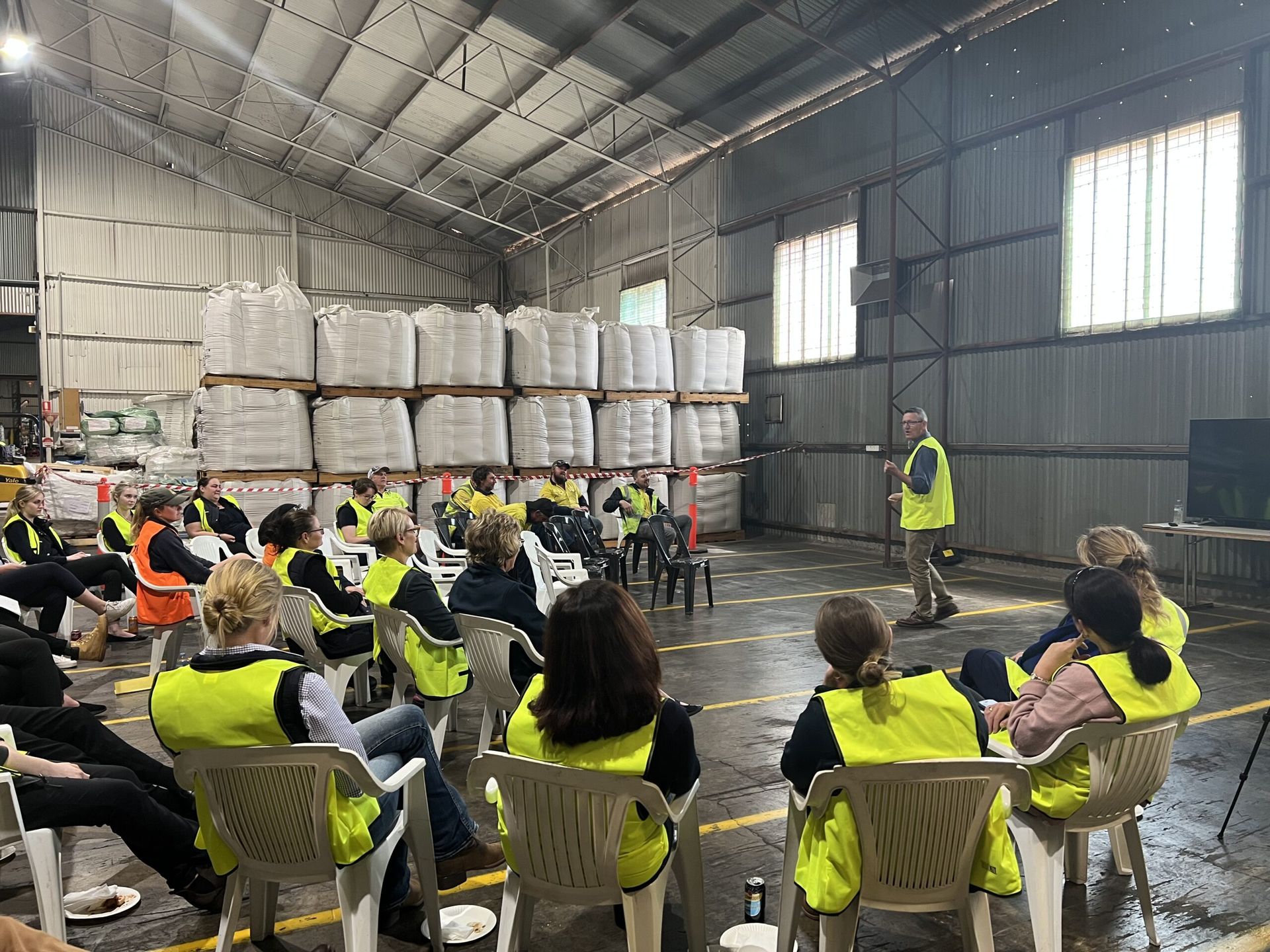 Warehouse safety briefing with seated workers in yellow vests and a presenter near stacked pallets and metal walls