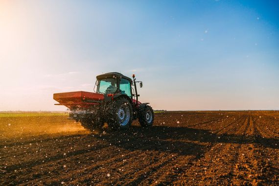 Tractor driving across a plowed field at sunset, spraying dust under a clear blue sky.