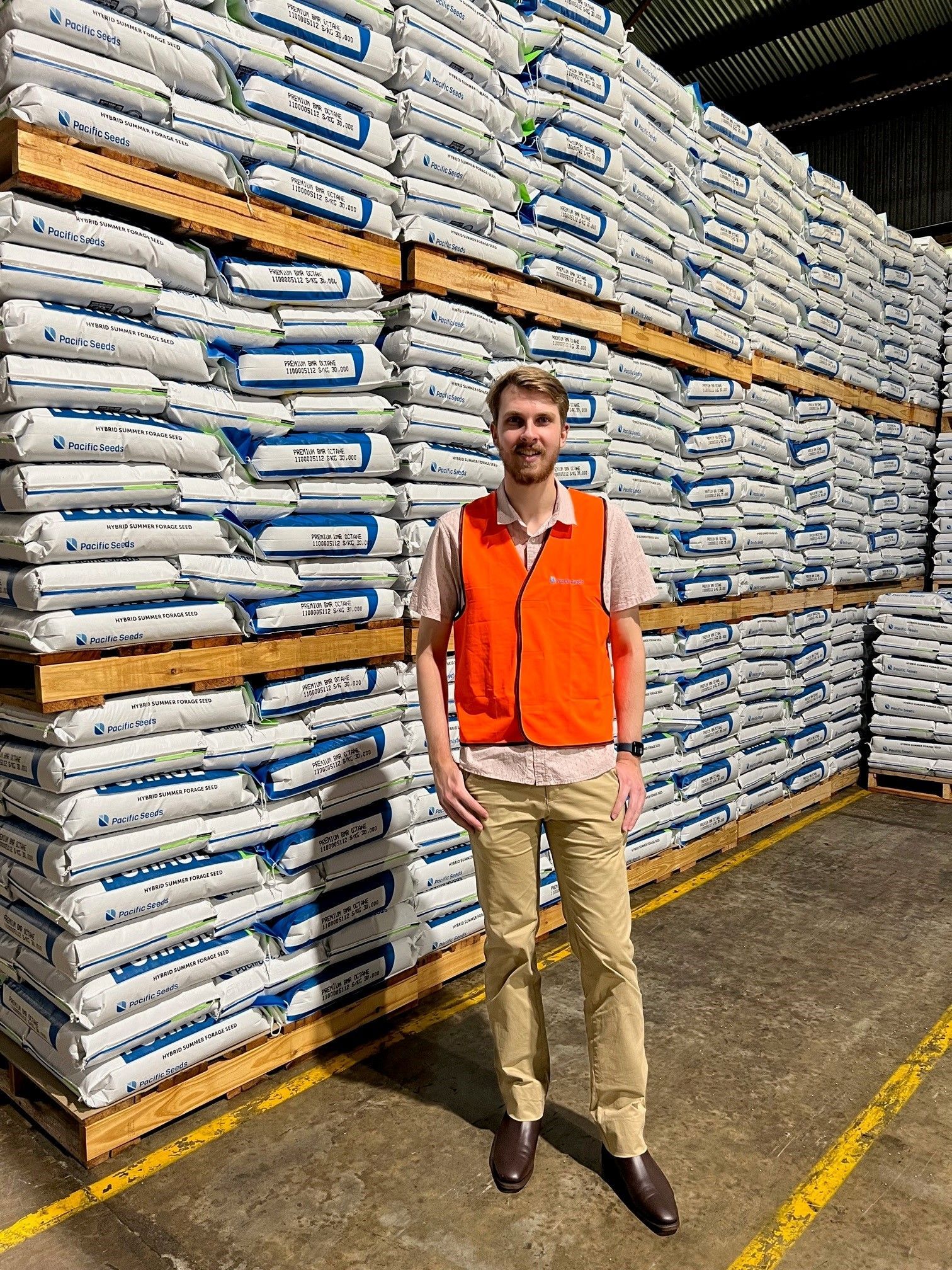 Man standing in a warehouse aisle beside stacked bags of goods, wearing an orange vest and khaki pants.