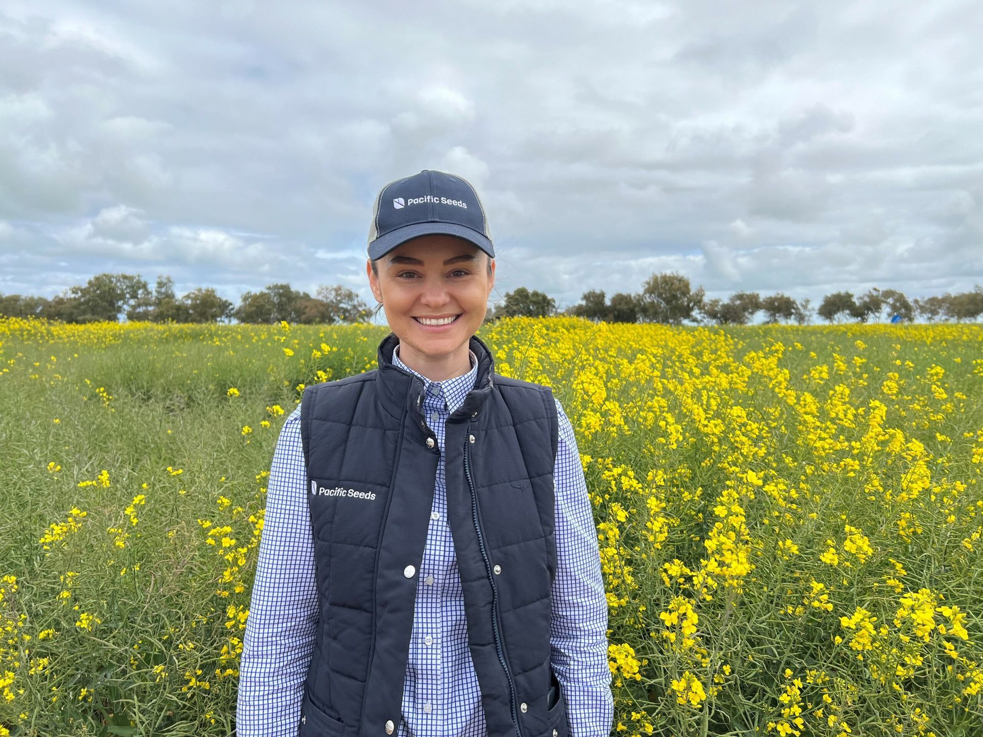 Smiling person in a cap and navy vest standing in a yellow wildflower field under a cloudy sky