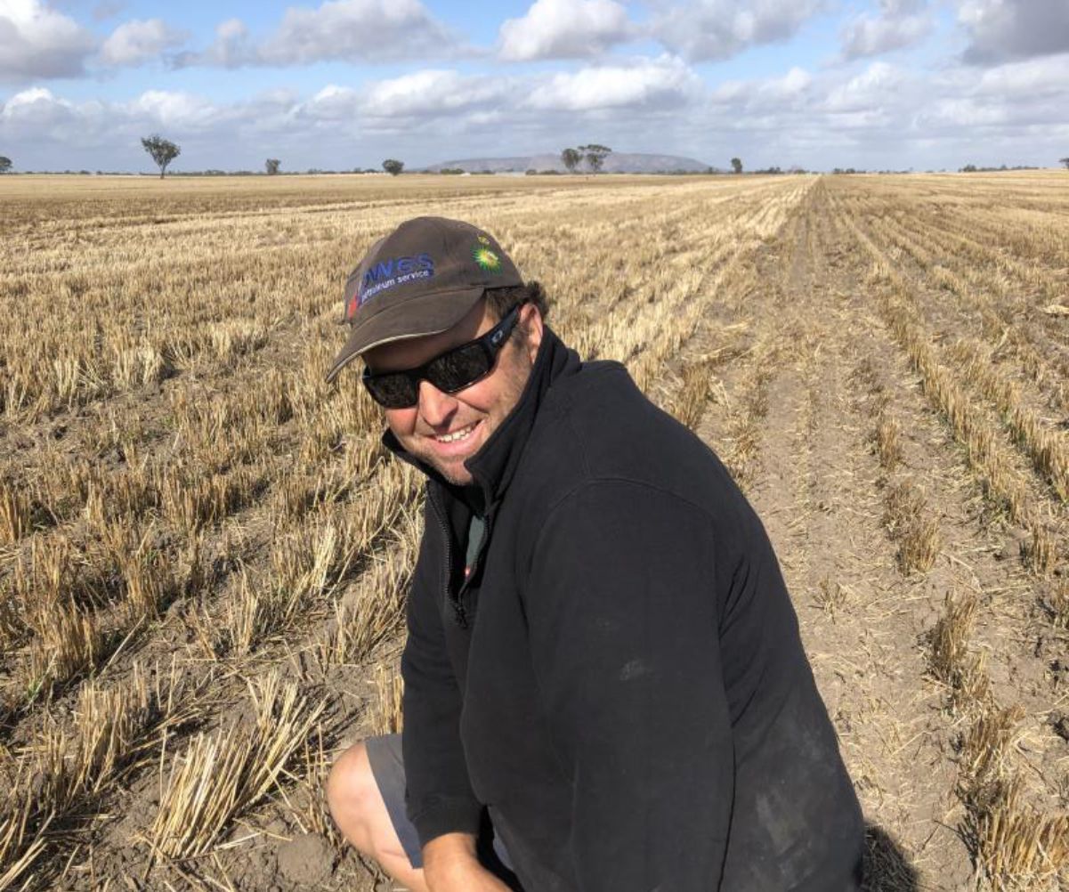 Smiling person in sunglasses and a cap kneels in a harvested field under a cloudy sky.