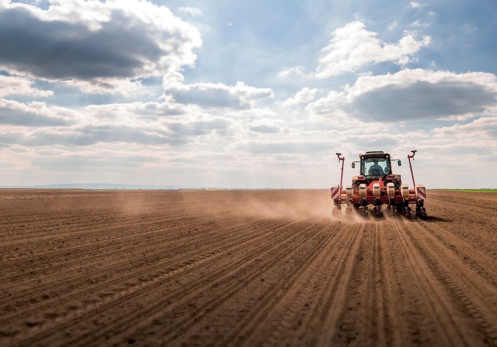 Red tractor tilling a wide brown field under a cloudy sky, kicking up dust