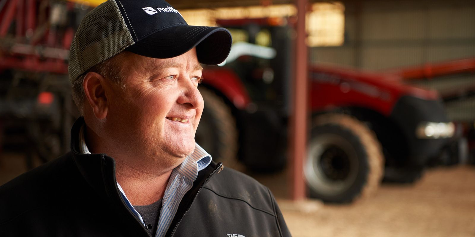 Man in a black cap and jacket smiling in a barn with a red tractor in the background