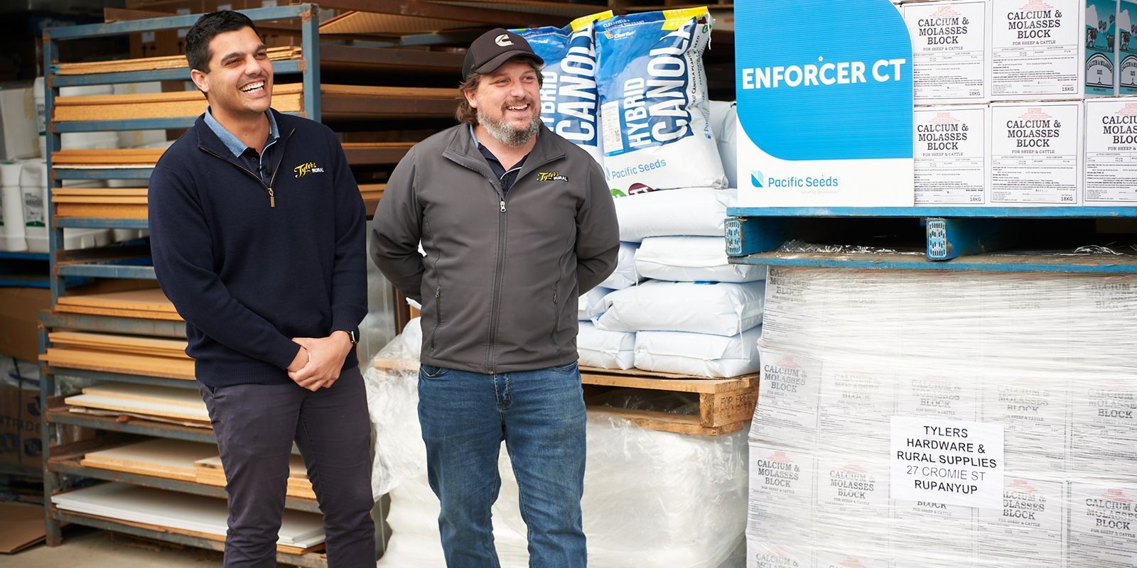 Two men standing beside stacked bags of Enforce CT products in a warehouse aisle.