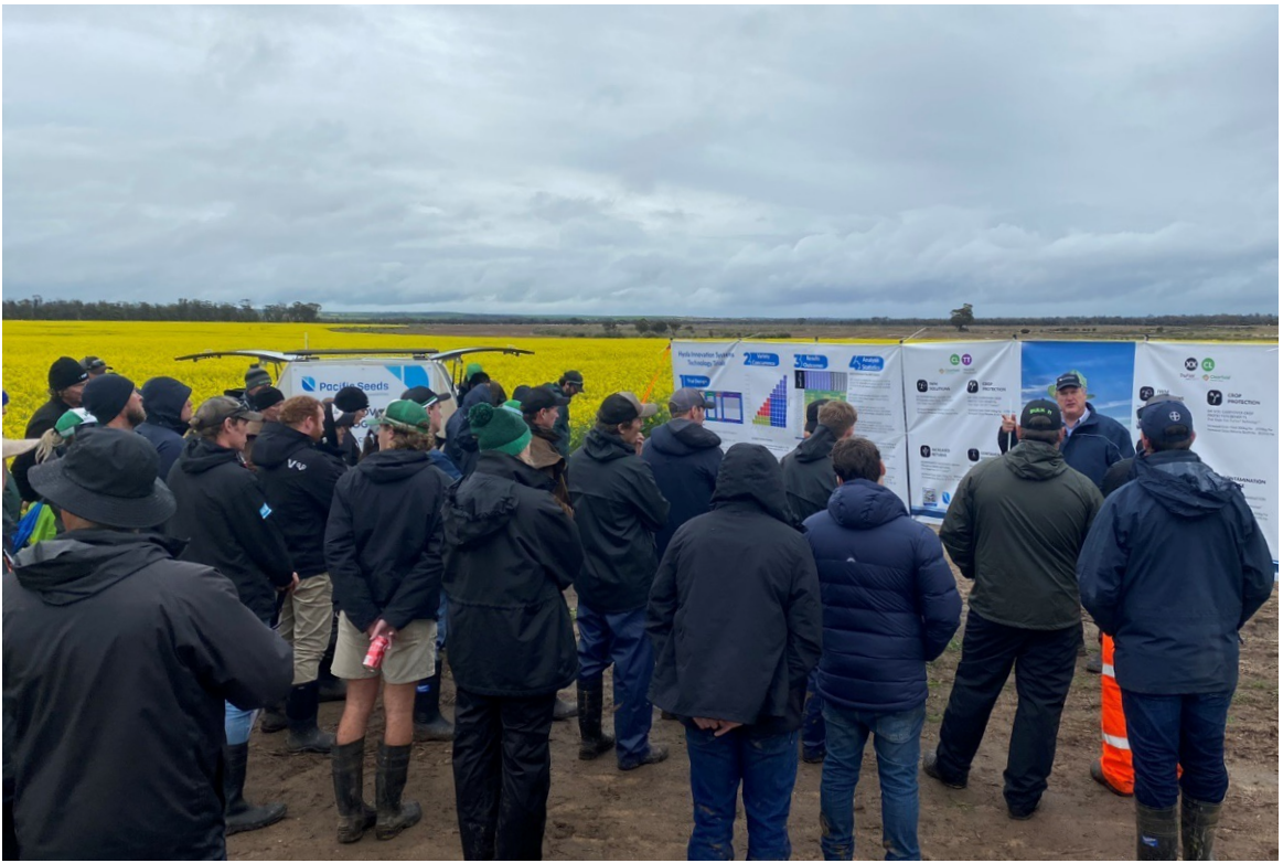 Crowd in dark coats watching a field demonstration beside large posters under a cloudy sky